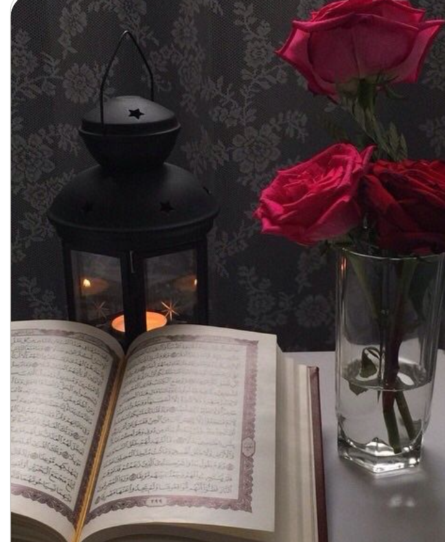 An open Quran beside a glowing lantern and a vase of roses in warm candlelight